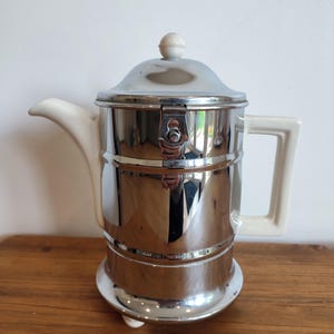May include: A silver and white ceramic coffee pot with a lid and a handle. The pot has a chrome finish and sits on three small feet.