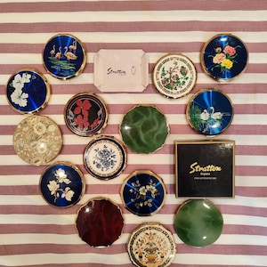Può includere: Collezione di specchietti compatti Stratton vintage con vari disegni, tra cui motivi floreali, di uccelli e geometrici. I compatti presentano bordi in metallo dorato e dettagli in smalto colorato. Alcuni compatti hanno uno sfondo blu. È presente anche una scatola Stratton.