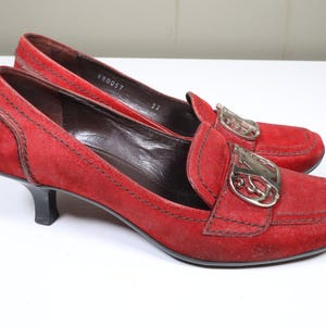 May include: A pair of red suede heeled loafers. The shoes have a silver-tone buckle detail on the vamp and a small, black heel. The shoes are size 37.