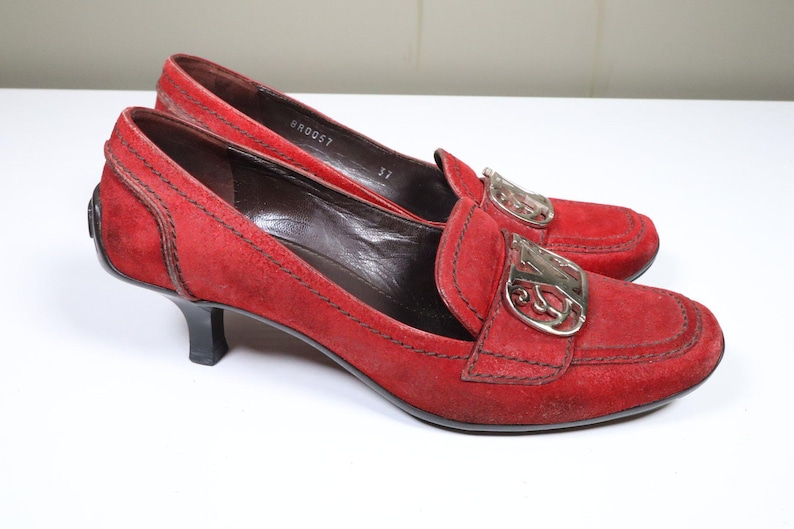 May include: A pair of red suede heeled loafers. The shoes have a silver-tone buckle detail on the vamp and a small, black heel. The shoes are size 37.