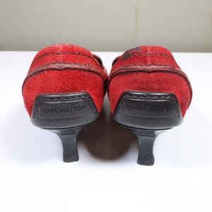 May include: A pair of red suede heeled shoes. The shoes have a rounded toe and a small heel. The heel is black and the sole is black. The brand name "LOUIS VUITTON" is printed on the heel.