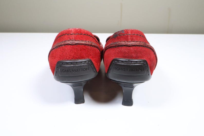 May include: A pair of red suede heeled shoes. The shoes have a rounded toe and a small heel. The heel is black and the sole is black. The brand name "LOUIS VUITTON" is printed on the heel.