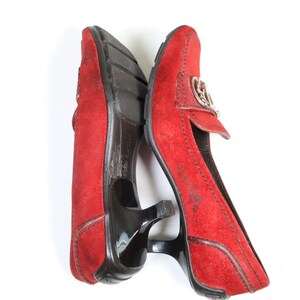 May include: Pair of red suede heeled loafers. The shoes have a pointed toe, a small heel, and a decorative silver-tone buckle. The soles are black. The shoes are displayed on a white background.