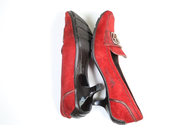 May include: Pair of red suede heeled loafers. The shoes have a pointed toe, a small heel, and a decorative silver-tone buckle. The soles are black. The shoes are displayed on a white background.