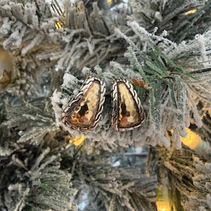 Pode incluir: Um par de brincos prateados e dourados. Os brincos têm um centro dourado martelado e uma borda prateada texturizada. São exibidos em um galho de pinheiro coberto de neve, sugerindo um tema de feriado.