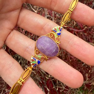 Pode incluir: Uma pulseira de ouro com uma grande pedra preciosa oval roxa. A pulseira tem elos de ouro detalhados e detalhes de esmalte coloridos em azul, verde, vermelho e branco. Pequenas pedras preciosas vermelhas são colocadas perto dos detalhes de esmalte.