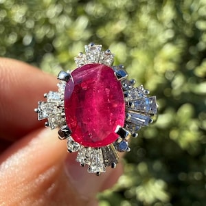 Puede incluir: Primer plano de un anillo con una gran gema de rubí de corte ovalado. El rubí es de color rojo vibrante y está rodeado de gemas más pequeñas y transparentes. El anillo tiene una banda plateada y un engaste detallado.