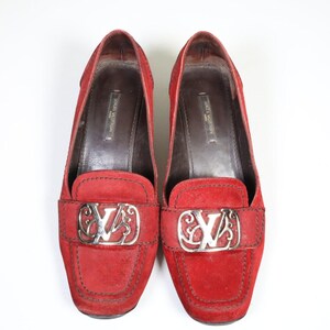 May include: Pair of red suede loafers with silver-tone hardware. The shoes feature a rounded toe and a decorative silver buckle with the brand's logo. The interior is a dark brown color.