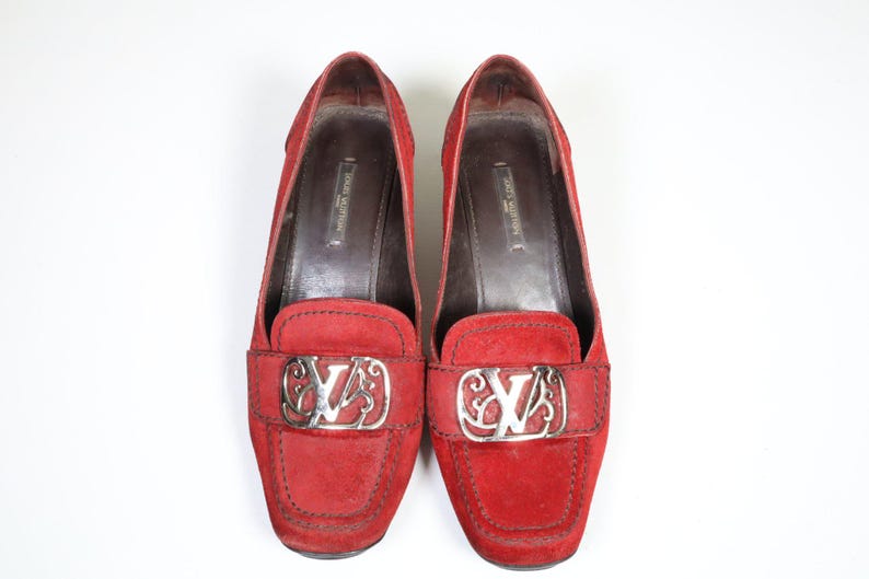 May include: Pair of red suede loafers with silver-tone hardware. The shoes feature a rounded toe and a decorative silver buckle with the brand's logo. The interior is a dark brown color.
