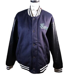 May include: Navy blue varsity jacket with black leather sleeves. The jacket features a white and black striped collar, cuffs, and hem. The word "Gucci" is embroidered in a light green script on the chest. The jacket has a button-up front.