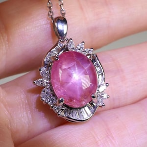 Könnte beinhalten: Eine silberne Anhängerkette mit einem großen, ovalen rosa Edelstein mit Sterneneffekt. Der Edelstein ist von kleineren, klaren Edelsteinen umgeben und in einen silbernen Metallrahmen gefasst. Die Halskette wird vor einem hellen Hintergrund gezeigt.