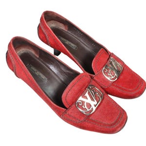 May include: A pair of red suede loafers with square toes. Each shoe features a silver-tone metal buckle with an ornate design and the initials "LV". The interior of the shoes is brown, and the heels are black.