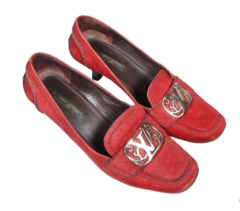 May include: A pair of red suede loafers with square toes. Each shoe features a silver-tone metal buckle with an ornate design and the initials "LV". The interior of the shoes is brown, and the heels are black.