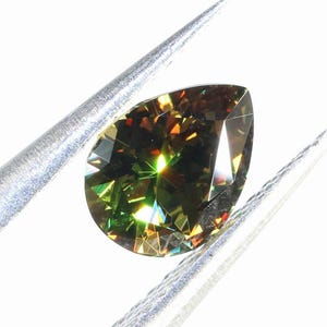 May include: A pear-shaped gemstone held by tweezers. The gem has a dark olive-green hue with flashes of yellow, orange, and green light. The facets of the stone reflect light, creating a sparkling effect against a white background.