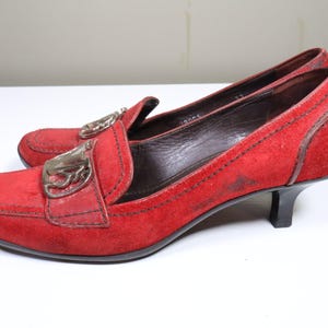 May include: A pair of red suede loafers with a low heel. The shoes have a decorative silver-tone buckle on the vamp and a stitched design around the edges. The heels are approximately 2 inches high.