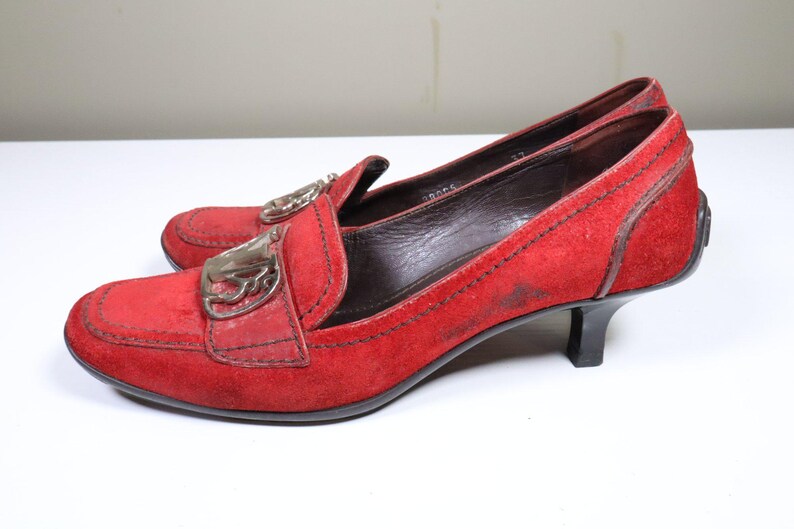 May include: A pair of red suede loafers with a low heel. The shoes have a decorative silver-tone buckle on the vamp and a stitched design around the edges. The heels are approximately 2 inches high.