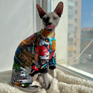 Peut inclure: Un chat Sphynx gris portant une veste colorée à motifs avec des graphismes de style bande dessinée. Le chat est assis sur une surface blanche et moelleuse près d'une fenêtre, les yeux fermés et la langue sortie.