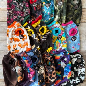 Könnte beinhalten: Eine Sammlung verschiedener Haustierpullover mit diversen Mustern, darunter Waschbären, Minecraft-Figuren, Tarnmuster, Corgis, Homer Simpson, Pferde und Pandas. Jedes Kleidungsstück hat einen Rundhalsausschnitt, Ärmelausschnitte und ein Markenetikett.