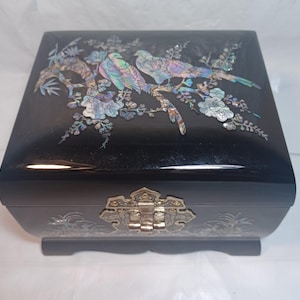 May include: A rectangular black lacquered jewelry box featuring iridescent mother-of-pearl inlay on the lid depicting two birds perched on flowering branches. The front includes a decorative silver-toned metal latch and additional floral inlay.