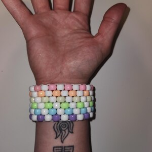 Puede incluir: Una pulsera de cuentas coloridas con filas de cuentas de colores pastel. La pulsera presenta cuentas rosas, blancas, naranjas, verdes, azules y moradas. La pulsera se usa en una muñeca con un tatuaje.