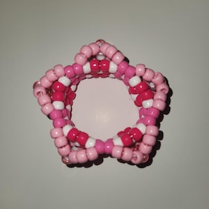 Puede incluir: Una pulsera en forma de estrella hecha de cuentas rosas y blancas. La pulsera está construida con pequeñas cuentas redondas, creando una superficie texturizada. El diseño presenta un efecto en capas, con las puntas de la estrella claramente definidas.