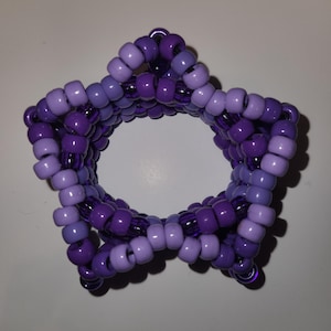 Puede incluir: Una pulsera en forma de estrella hecha de cuentas moradas. La pulsera presenta un degradado de tonos morados, desde lavanda claro hasta violeta intenso. Las cuentas están ensartadas para formar un diseño de estrella de cinco puntas, creando un accesorio único.
