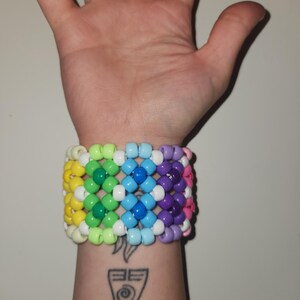Puede incluir: Una colorida pulsera de cuentas con un patrón de arcoíris, con cuentas amarillas, verdes, azules, moradas y rosas. La pulsera se lleva en una muñeca con un tatuaje de dos símbolos.