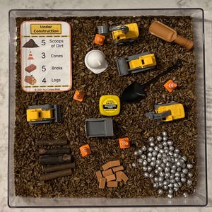 May include: A sensory bin with a brown, shredded paper-like material.  The bin contains toy construction vehicles, logs, bricks, cones, and a measuring tape.  A card with the text "Under Construction" lists the number of each item in the bin.