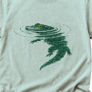 Bay Comfort Colors shirt featuring a vintage-style green alligator partially submerged in water with circular ripples and a bright yellow-green eye.