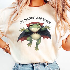 May include: Ivory t-shirt with a cartoon frog dressed as a vampire, wearing a black cape and red collar. The text "OFF TO COMMIT JUMP SCARES" is printed above the frog. The shirt is made of a soft material.