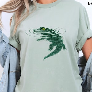 May include: A pale green t-shirt with a green illustration of an alligator in water. The alligator's eye is bright green. The shirt has rolled-up sleeves. The word "BAY" is printed in black at the top right.