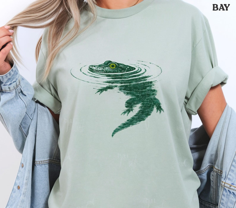 May include: A pale green t-shirt with a green illustration of an alligator in water. The alligator's eye is bright green. The shirt has rolled-up sleeves. The word "BAY" is printed in black at the top right.
