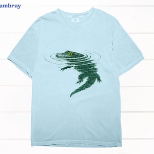 May include: Pale blue t-shirt featuring a green alligator design. The alligator is shown in water, with concentric circles around its head. The word "chambray" is in the upper left corner. A casual wear item.