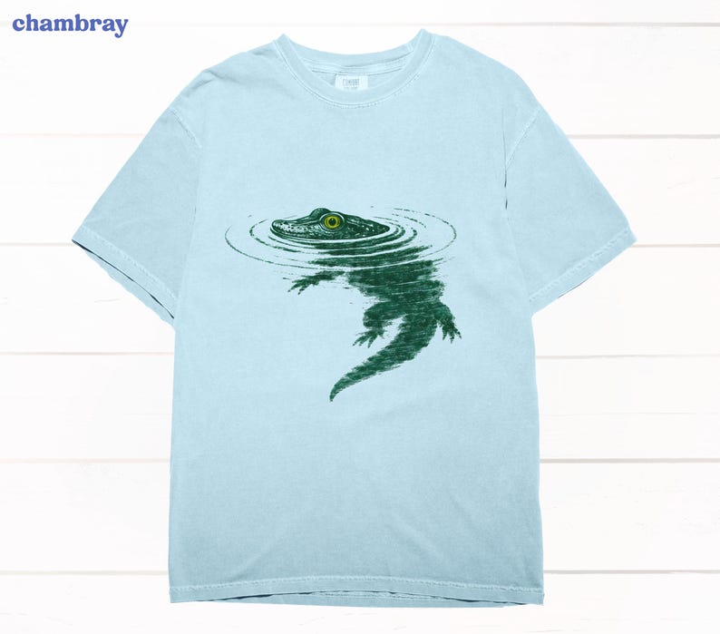 May include: Pale blue t-shirt featuring a green alligator design. The alligator is shown in water, with concentric circles around its head. The word "chambray" is in the upper left corner. A casual wear item.
