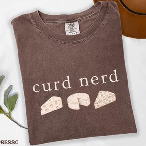 May include: Espresso-coloured t-shirt featuring the words "curd nerd" and illustrations of three cheeses. The shirt has a crew neck and is made of a soft material. The Comfort Colors label is visible.