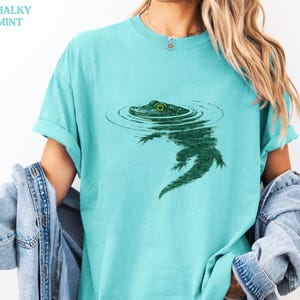 May include: A light teal t-shirt with a green alligator graphic in water. The alligator has a detailed design with a visible eye. The text "CHALKY MINT" is in the upper left corner. The shirt has rolled-up sleeves.