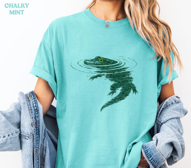May include: A light teal t-shirt with a green alligator graphic in water. The alligator has a detailed design with a visible eye. The text "CHALKY MINT" is in the upper left corner. The shirt has rolled-up sleeves.