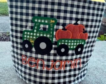 May include: A black and white buffalo check fabric bucket with a green tractor and orange pumpkin applique. The name Benjamin is embroidered in orange thread below the tractor. The bucket features a handle and a white label at the top.