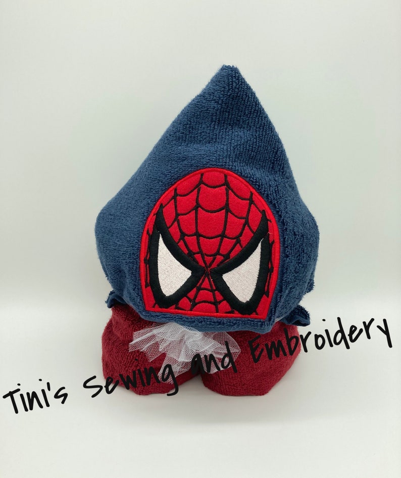 May include: A blue hooded towel with a red and black embroidered Spiderman face on the hood. The towel is rolled up and tied with a white ribbon. The text "Tini's Sewing and Embroidery" is visible in the bottom of the image.