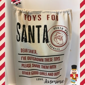 May include: A white canvas bag with a drawstring top. The bag has black text that reads "TOYS FOR SANTA" and "DEAR SANTA, I'VE OUTGROWN THESE TOYS PLEASE SHARE THEM WITH OTHER GOOD GIRLS AND BOYS LOVE. Jasmine". There is a red and white striped border around the image.