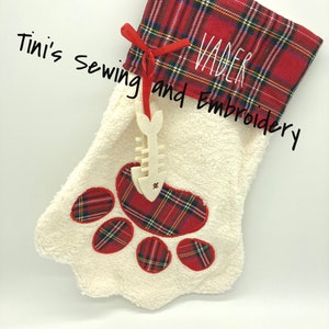 May include: Red and white plaid Christmas stocking with a white paw print design and the name "Vader" embroidered on the cuff. The stocking is decorated with a white wooden fish bone ornament. The text "Tini's Sewing and Embroidery" and "Farmhouse Font" are visible below the stocking.