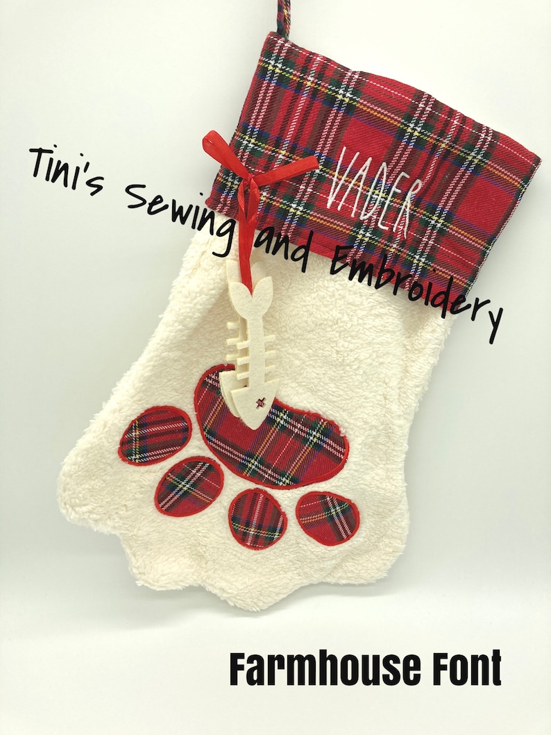May include: Red and white plaid Christmas stocking with a white paw print design and the name "Vader" embroidered on the cuff. The stocking is decorated with a white wooden fish bone ornament. The text "Tini's Sewing and Embroidery" and "Farmhouse Font" are visible below the stocking.