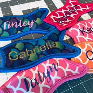 May include: Five personalized mermaid tail can coolers in various colors. The coolers are made of neoprene and have a mermaid scale pattern. The names Finley, Gabriella, Madison, Falyyn, and Ken are embroidered on the coolers.