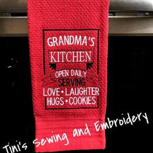 May include: Red kitchen towel with black embroidered text that reads "Grandma's Kitchen Open Daily Serving Love Laughter Hugs Cookies". The towel is hanging on a silver oven door handle. The text "Tini's Sewing and Embroidery" is handwritten in white on the bottom of the towel.