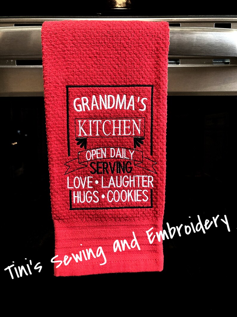 May include: Red kitchen towel with black embroidered text that reads "Grandma's Kitchen Open Daily Serving Love Laughter Hugs Cookies". The towel is hanging on a silver oven door handle. The text "Tini's Sewing and Embroidery" is handwritten in white on the bottom of the towel.