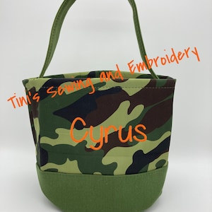 May include: A green camouflage fabric bucket bag with a green handle. The bag has the name "Cyrus" embroidered on the front in orange thread. The text "Tini's Sewing and Embroidery" is written in orange text at the top of the image. The text "Friendship Font" is written in black text at the bottom of the image.