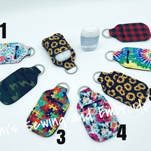 K&ouml;nnte beinhalten: Zehn verschiedene Handdesinfektionsmittel-Halter mit Schl&uuml;sselanh&auml;ngern. Die Halter sind aus Stoff mit verschiedenen Mustern, darunter Blumen, Tarnmuster, Puzzle-Teile, Sonnenblumen und Batik. Die Zahlen 1 bis 7 sind auf dem Bild geschrieben.