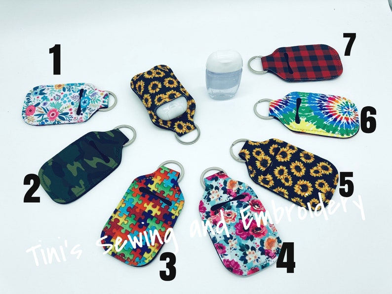 K&ouml;nnte beinhalten: Zehn verschiedene Handdesinfektionsmittel-Halter mit Schl&uuml;sselanh&auml;ngern. Die Halter sind aus Stoff mit verschiedenen Mustern, darunter Blumen, Tarnmuster, Puzzle-Teile, Sonnenblumen und Batik. Die Zahlen 1 bis 7 sind auf dem Bild geschrieben.
