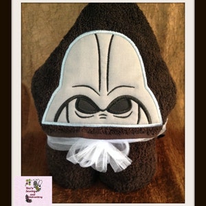 May include: A brown hooded towel with a white bow and an embroidered Darth Vader face on the hood. The towel is folded and displayed on a brown surface.