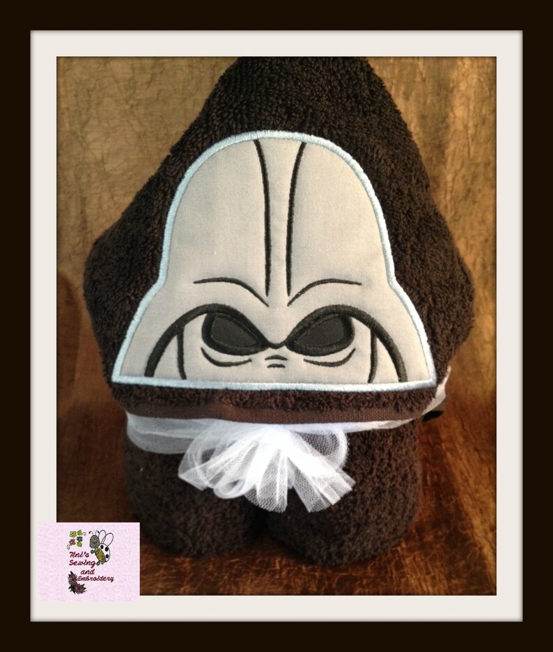 May include: A brown hooded towel with a white bow and an embroidered Darth Vader face on the hood. The towel is folded and displayed on a brown surface.